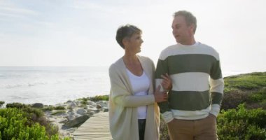 Kafkasyalı çift, güneşli bir günde deniz kenarında ağır çekimde yürümenin keyfini çıkarıyorlar. Sahilde erken emeklilik.