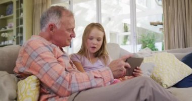 Kafkasyalı kıdemli bir adam ve torunu oturma odasındaki kanepede oturup ağır çekimde tablet bilgisayar kullanıyorlar..