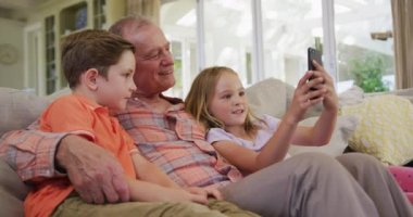 Kafkasyalı kıdemli bir adam, torunu ve torunu evde birlikte vakit geçiriyorlar, oturma odasında bir kanepede oturuyorlar, akıllı telefon kullanıyorlar, ağır çekimde..