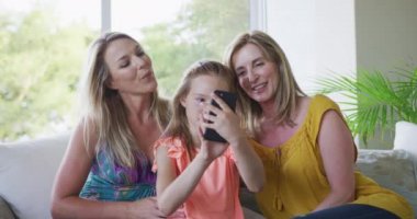 Kıdemli beyaz kadın, kızı ve torunu, evde birlikte vakit geçiriyorlar, oturma odasında bir kanepede oturuyorlar, akıllı telefon kullanıyorlar ve gülümsüyorlar, ağır çekimde..