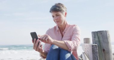 Kafkasyalı bir kadın güneşli bir günde deniz kenarında boş vakit geçirmenin keyfini yavaş çekimde akıllı telefon kullanarak sürüyor. Sahilde erken emeklilik.