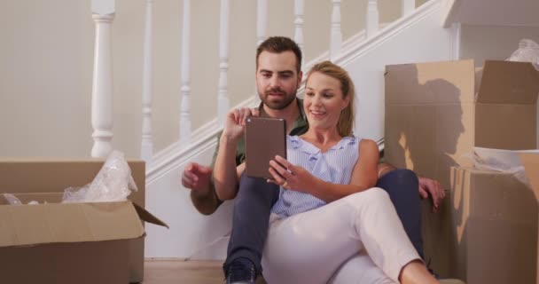 白种人夫妇搬进新屋 坐在地上 使用平板电脑 微笑着 四周都是纸板箱 动作缓慢 — 图库视频影像