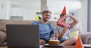 Beyaz bir adam oğluyla birlikte vakit geçiriyor, bir kanepede oturuyor, oğlunun doğum gününü kutluyor, bir hediye açıyor, dizüstü bilgisayar kullanıyor. Covid 19 koronavirüs karantina süresince sosyal mesafeler.