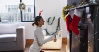 Noel 'de evde beyaz bir kadın, ren geyiği boynuzu takıyor, oturma odasında yerde oturuyor, şömineyi dekore ediyor, ağır çekimde. Covid 19 Coronavirus karantina süresince sosyal mesafeler.