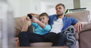 Beyaz bir adam oğluyla birlikte vakit geçiriyor, kanepede oturuyor, televizyon seyrediyor. Covid 19 koronavirüs karantina süresince sosyal mesafeler.