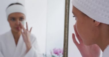 Beyaz kadın evde vakit geçiriyor, banyoda dikiliyor, aynaya bakıyor, yüz maskesi takıyor, ağır çekimde. Covid 19 Coronavirus karantina süresince sosyal mesafeler.