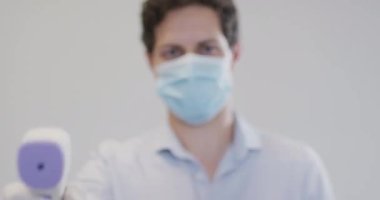 Beyaz bir adam, yüz maskesi ve eldiven takmış, sıcaklığı dijital termometreyle yavaş çekimde kontrol ediyor. Coronavirus Covid 19 salgını sırasında işyerinde sağlık ve hijyen.