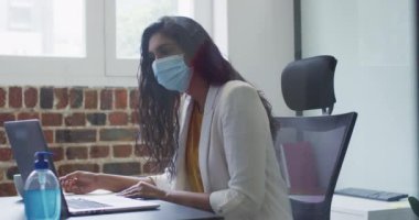 Karışık ırk kadın iş yaratıcılığı, modern ofiste maske takmak, dizüstü bilgisayarı yavaş çekimde kullanmak. Coronavirus Covid 19 salgını sırasında işyerinde sağlık ve hijyen.
