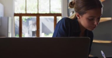 Kafkasyalı kadın mutfakta oturuyor, dizüstü bilgisayar kullanıyor ve kalemle notlar yazıyor, ağır çekimde. Covid 19 Coronavirus karantina süresince sosyal mesafeler.