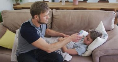 Beyaz bir adam oğluyla kanepede oturup vakit geçiriyor. Babası oğlunun ateşini ölçüyor. Covid 19 koronavirüs karantina süresince sosyal mesafeler.