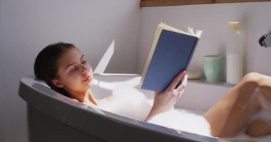 Beyaz kadın banyoda zaman geçiriyor, kitap okuyup rahatlıyor, gülümsüyor, ağır çekimde. Covid 19 Coronavirus karantina süresince sosyal mesafeler.