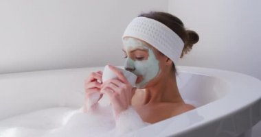 Beyaz kadın banyoda vakit geçiriyor, yüz maskesi takıyor, banyoda dinleniyor, kahve içiyor, ağır çekimde. Covid 19 Coronavirus karantina süresince sosyal mesafeler.
