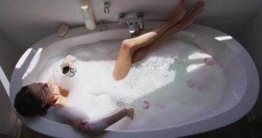 Beyaz bir kadının banyoda, ayaklarını kaldırmış, çay içerek ve ağır çekimde banyoda vakit geçirmesi. Covid 19 Coronavirus karantina süresince sosyal mesafeler.