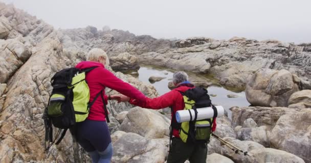 山の中で岩を登りながら バックパックとトレッキングポールを手にしたシニアハイカーカップル トレッキング ハイキング アクティビティ 冒険の概念 — ストック動画
