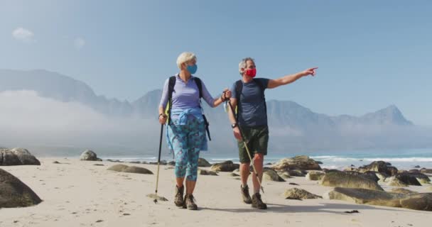 年长的登山者夫妇戴着面罩 背着背包和远足杆 一边在海滩上远足 一边指明方向 冒险的概念 冒险的概念 — 图库视频影像