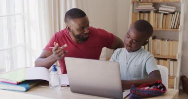 Evde dizüstü bilgisayar ve dezenfektan şişesiyle otururken Afrikalı Amerikalı baba oğluna ödevlerinde yardım ediyor. Covid 19 koronavirüs karantina süresince sosyal mesafeler.