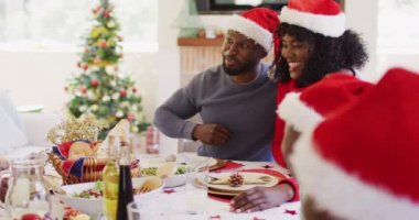 Noel Baba şapkalı Afro-Amerikan çift evde birlikte yemek yerken gülümseyip kadeh kaldırıyor. Noel kutlaması geleneği konsepti