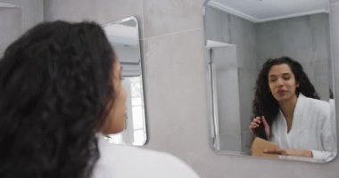 Banyoda saçını tarayan, bornoz giyen ve aynaya yavaş çekimde bakan mutlu melez kadın. Coronavirus covid 19 tecridi sırasında evde kaliteli zaman geçirmenin keyfini çıkarıyorum..