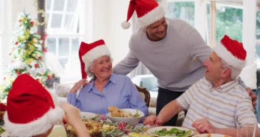 Noel Baba şapkalı beyaz çift evdeki yemek masasında otururken oğullarını kucaklıyor ve birlikte öğle yemeğinin tadını çıkarıyor. Noel kutlaması geleneği konsepti