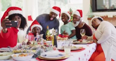 Noel Baba şapkalı Afro-Amerikan aile evde birlikte yemek yerken akıllı telefondan selfie çekiyor. Noel kutlaması geleneği konsepti