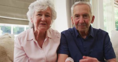 Kameraya gülümseyen ve el sallayan yaşlı beyaz bir çiftin portresi oturma odasında ağır çekimde oturuyor. Coronavirus covid 19 tecridi sırasında evde vakit geçirmenin tadını çıkarıyor..