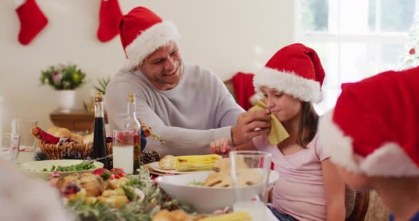 白种人老人头戴圣诞礼帽 用餐巾纸擦拭女儿的脸 坐在饭桌上 一起在家里吃午饭 圣诞节庆祝传统的概念 — 图库视频影像