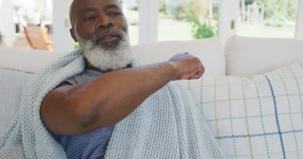アフリカ系アメリカ人の男性が顔を肘で覆い ゆっくりとした動きでリビングルームのソファの上に座って くしゃみをします コロナウイルスCovid 19のロックダウン中に自宅で時間を楽しむ — ストック動画