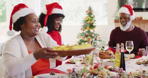 Africká seniorka v Santa klobouku podávala jídlo svému synovi a vnukovi, zatímco seděli na jídelním stole a obědvali spolu doma. vánoční slavnostní tradice koncepce oslav