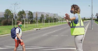 Yüz maskesi takan beyaz bir bayan öğretmen ve yüksek görüş açısına sahip bir yelek yaya geçidinde trafiği durduruyor böylece okul maskeli çocuklar yolu güvenle geçebiliyor. Kovid19 Coronavirus salgını sırasında yol güvenliği eğitimi.