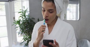 Karışık ırk kadını dişlerini fırçalıyor ve banyoda akıllı telefonunu kullanıyor. Kafasına havlu sarılı bornoz giyiyor. Ağır çekimde. Coronavirus covid 19 tecridi sırasında evde kaliteli zaman geçirmenin keyfini çıkarıyorum..