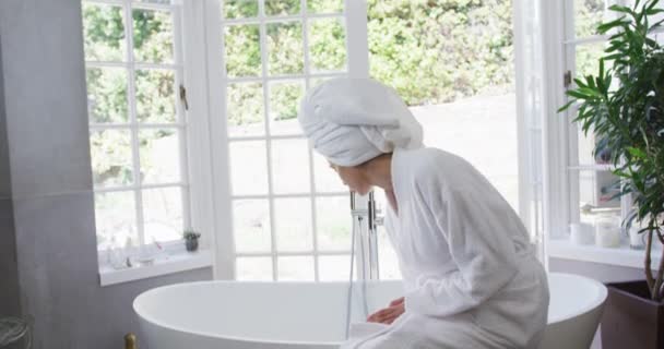 混合赛女子检查水 坐在浴室的浴缸上 身穿浴衣 头上裹着毛巾 动作缓慢 在科罗纳韦病毒入侵期间 在家中享受美好的时光 — 图库视频影像