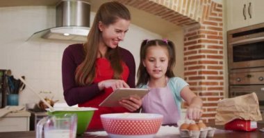 Beyaz anne ve kız önlük giyip evdeki mutfakta yemek pişirirken dijital tablet kullanıyorlar. Koronavirüs salgını sırasında karantina karantinasına sosyal uzaklık