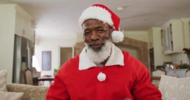 Noel zamanı Noel Baba kostümü giyen, hediyeleri gösteren ve kameraya el sallayan, oturma odasında ağır çekimde koltukta oturan mutlu Afrikalı Amerikalı adamın portresi. Coronavirus covid 19 'un tecridi sırasında evde geçirilen Noel' in tadını çıkarıyorum..