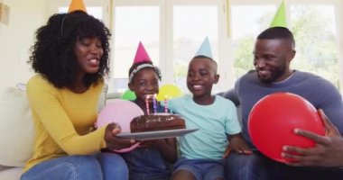 Afrikalı Amerikalı çocuk pastaya mum üflüyor ve doğum gününü ailesiyle evde kutluyor. Coronavirus karantinası süresince doğum günü kutlaması sosyal mesafesi