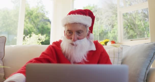 サンタの衣装を着て プレゼントを見せ ビデオチャットのためにラップトップコンピュータを使用して 遅い動きでリビングルームのソファに座って クリスマス時にシニア白人男性 コロナウイルスCovid 19ロックダウン中に自宅でクリスマスを楽しむ — ストック動画