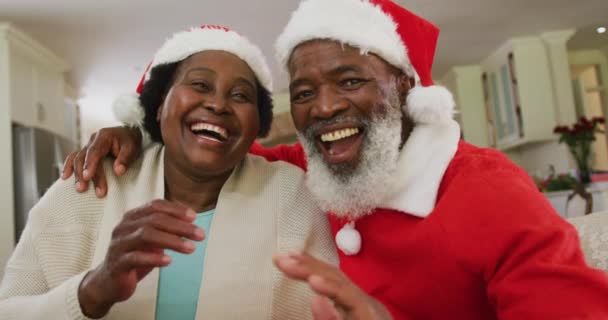 クリスマスにサンタの帽子をかぶったアフリカ系アメリカ人夫婦の肖像画 サンタの衣装を着た男 手を振ってカメラにキスを吹いて スローモーションでリビングルームのソファに座っています コロナウイルスCovid 19のクリスマスの時間を自宅で楽しむ — ストック動画