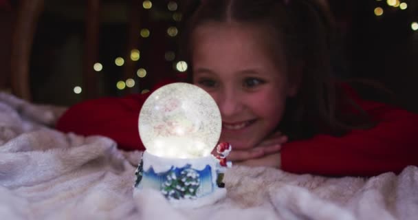Blank Meisje Glimlachend Kijkend Naar Sneeuwbol Terwijl Liggend Onder Deken — Stockvideo