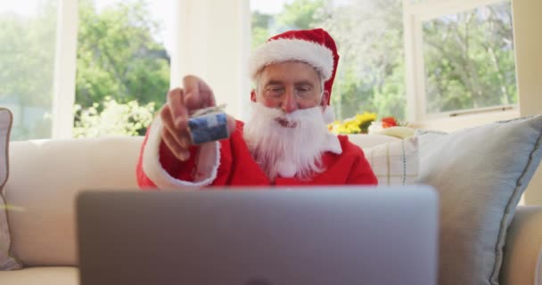サンタの衣装を着てクリスマスの時間にシニア白人男性は ビデオチャットのためのラップトップコンピュータを使用して スローモーションでリビングルームのソファに座って 現在を示しています コロナウイルスCovid 19ロックダウン中に自宅でクリスマスを楽しむ — ストック動画