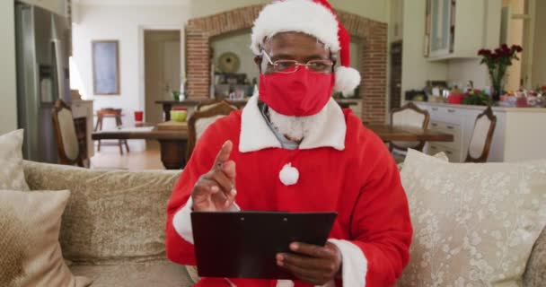 上了年纪的非洲裔美国人 圣诞节时戴着红脸面具 身穿桑塔服装 写着和拿着剪贴板 坐在客厅的沙发上慢动作 — 图库视频影像