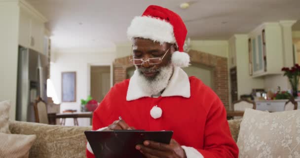 クリスマスの時間にサンタの衣装を着たシニアアフリカ系アメリカ人の男性は ゆっくりとした動きでリビングルームのソファに座って クリップボードを書き 保持します コロナウイルスCovid 19ロックダウン中に自宅でクリスマスを楽しむ — ストック動画