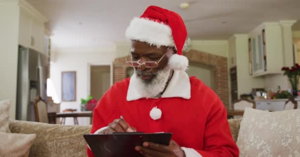 クリスマスの時間にサンタの衣装を着たシニアアフリカ系アメリカ人の男性は ゆっくりとした動きでリビングルームのソファに座って クリップボードを書き 保持します コロナウイルスCovid 19ロックダウン中に自宅でクリスマスを楽しむ — ストック動画