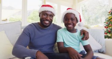 Afrikalı Amerikalı baba ve oğul kameraya bakıyor. Evde Noel 'de pazılarını esnetip el sallıyorlar. Coronavirus karantinası sırasındaki sosyal mesafeyi kutluyoruz.