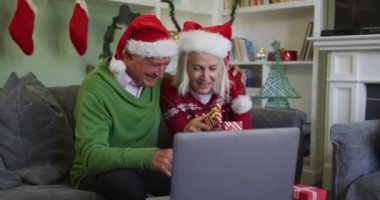 Mutlu Kafkasyalı çift Noel 'i Noel Baba şapkası takarak kutluyor, dizüstü bilgisayarla video görüşmesi yapıyor, hediyeler tutuyor, kanepede ağır çekimde oturuyor. Coronavirus covid 19 salgını sırasında evde birlikte vakit geçirmek..