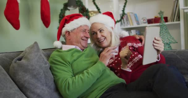 快乐的老加卡人夫妇戴着圣诞礼帽庆祝圣诞 用平板电脑打一个视频电话 坐在沙发上慢吞吞地坐在家里 食管癌大流行期间在家共度的美好时光 — 图库视频影像