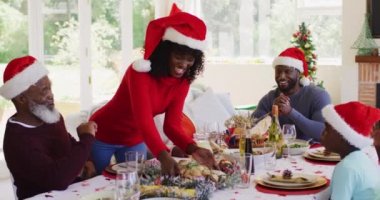 Afrika kökenli Amerikalı aile Noel Baba şapkası takıyor. Annem, Noel 'de evde öğle yemeği yemeden önce yemek masasında oturan aileye yemek servisi yapıyor. Noel bayramı geleneği kutlaması