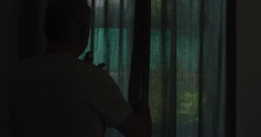 Evde, pencerenin önünde dikiz aynasından bakan yaşlı beyaz adam perdeleri indiriyor ve yavaş çekimde dışarı bakıyor. Coronavirus covid 19 salgını sırasında evde..