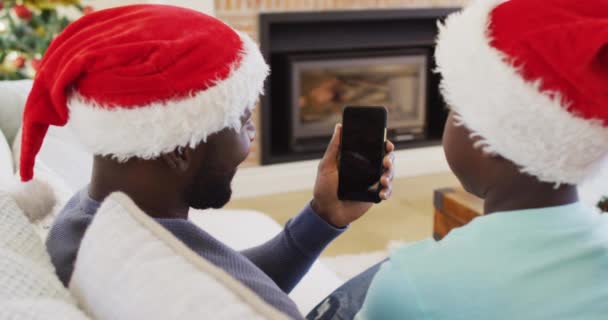 Afrikai Amerikai Apa Fia Videokazettával Okostelefonon Télapó Sapkát Visel Karácsonykor — Stock videók