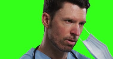 Beyaz erkek doktor yeşil ekran arka planında yüz maskesi takıyor. Adam ameliyat önlüğü takıyor, maskesini çıkarıyor. Coronavirus covid 19 salgını sırasında tıp alanında hijyen..