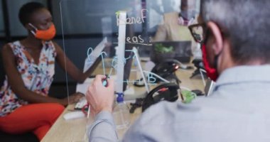 Modern ofisteki masasında otururken cam tahtaya yazı yazan yüz maskesi takan bir adamın dikiz görüntüsü. Koronavirüs salgını sırasında karantinaya alınma