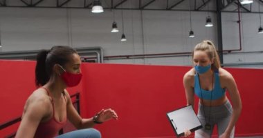 Spor salonunda yüz maskesi takmış, beyaz kadın zamanlaması yapan karışık ırklı bir kadın. Coronavirus covid 19 salgını sırasında spor yaparken spor salonunda hijyen..